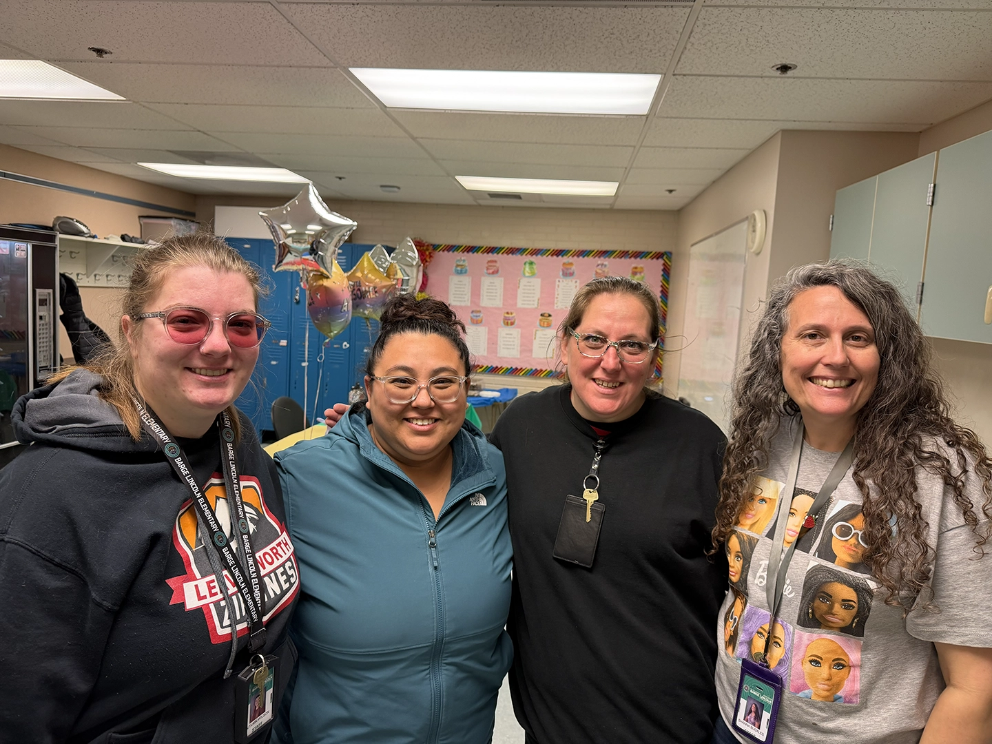 Four women smiling and standing side by side in an office, each holding a small gift box with a colorful Valentine’s Day label, wearing matching dark gray “Communication looks different for everyone” t-shirts.