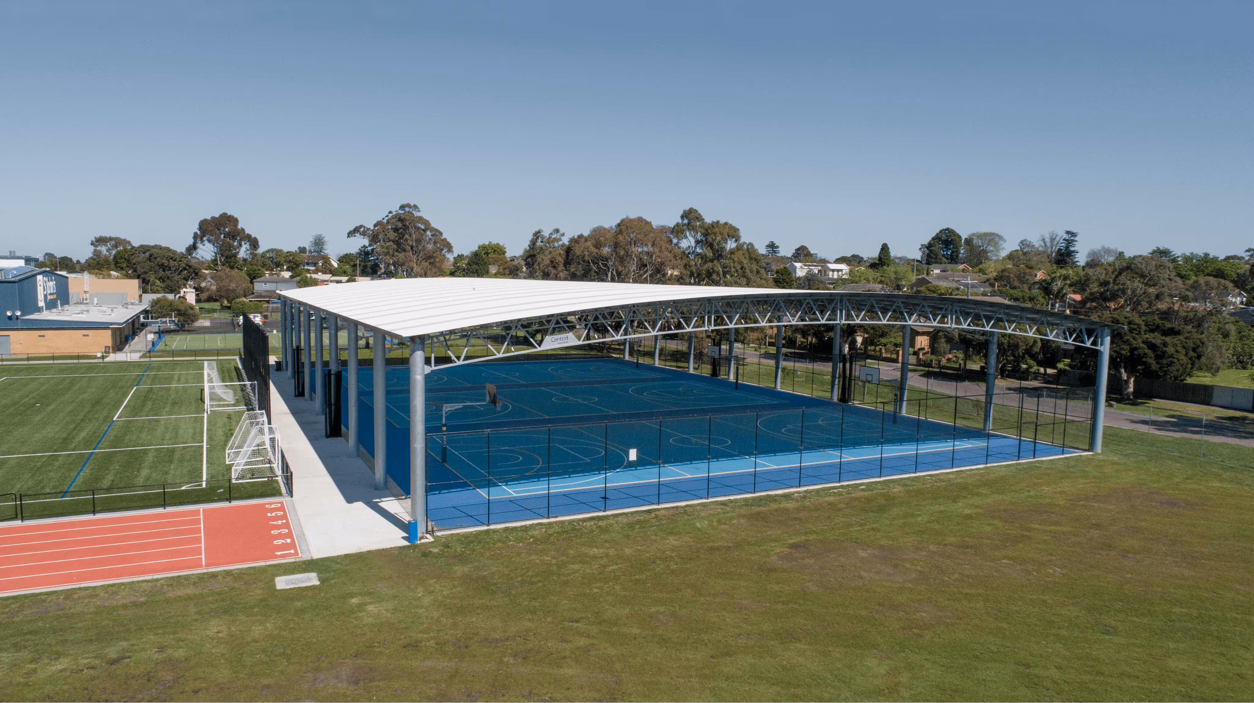 Outdoor sports complex with a covered blue basketball court, adjacent green soccer field, and red running track under a clear sky.