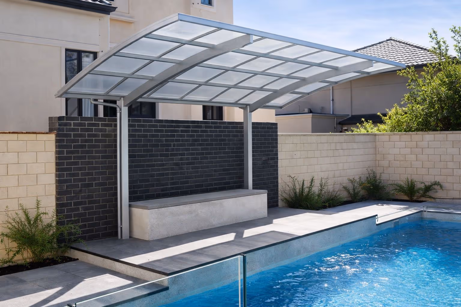 Modern outdoor poolside seating area with a sleek glass and metal cantilevered canopy and a concrete bench against a dark brick wall.