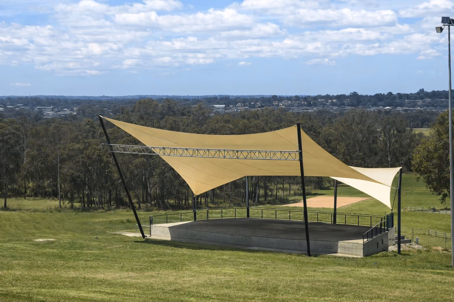 Outdoor stage with beige sail shade canopy set on a grassy hillside with forest and cityscape in the background.