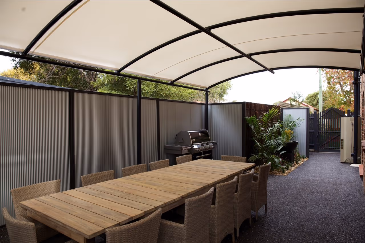 Outdoor dining area with a long wooden table surrounded by wicker chairs under a curved canopy, next to a barbecue grill and decorative plants.