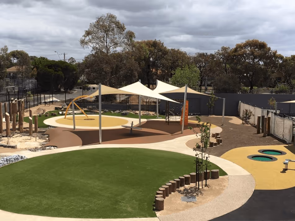 Playground with shaded canopies, artificial turf, wooden stepping logs, and play equipment under a cloudy sky.