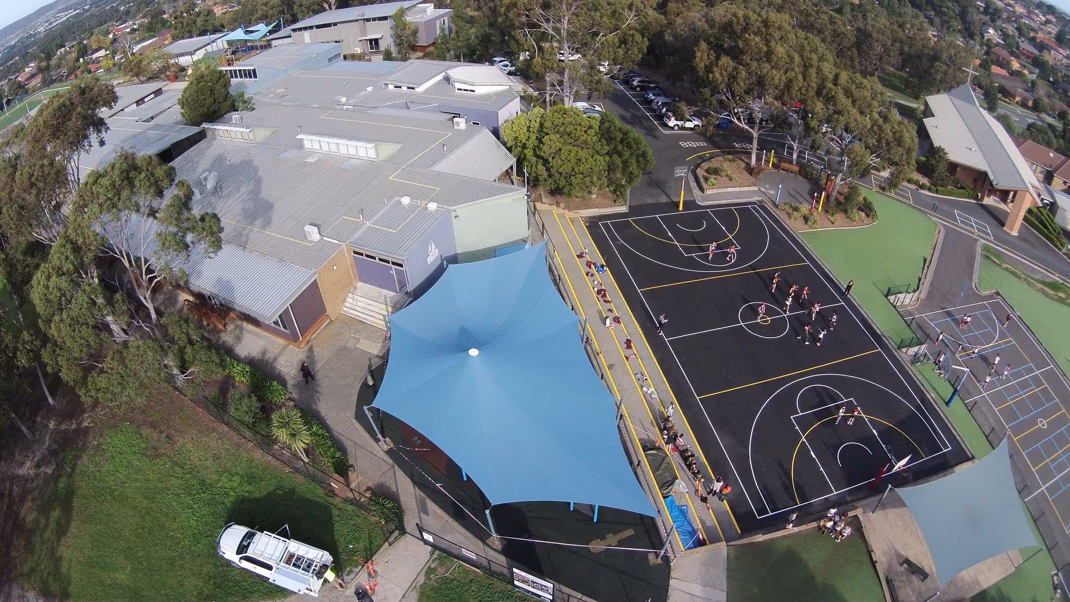 Aerial view of a school sports area featuring a large blue shade sail covering a playground and adjacent basketball courts with children playing.
