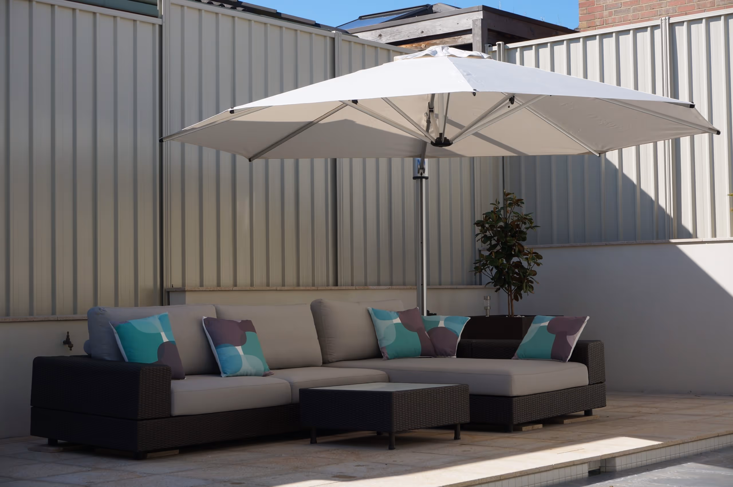 Outdoor patio scene with a black wicker sectional sofa with gray cushions and colorful pillows under a large white umbrella.