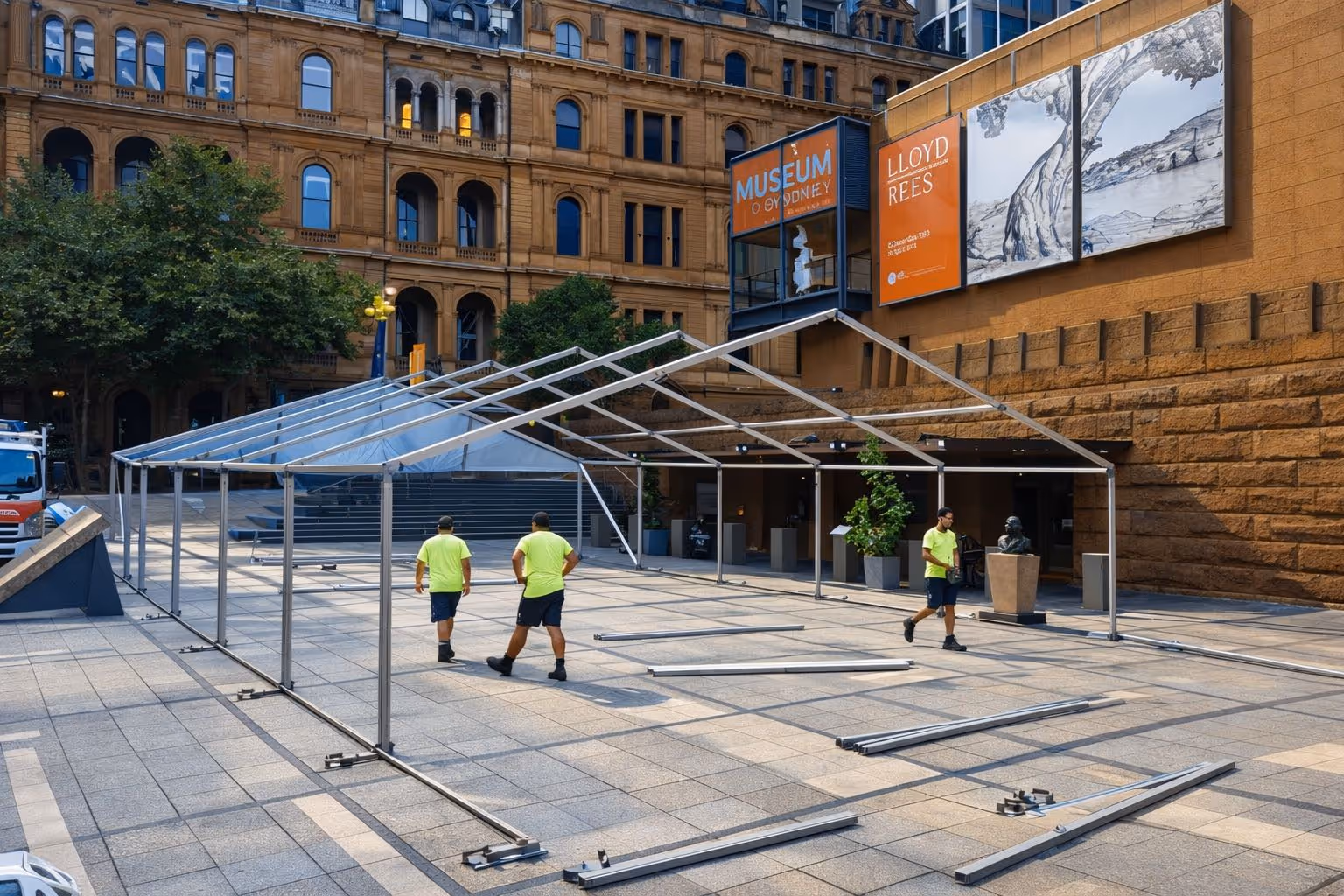 Three workers in yellow shirts assembling a large metal frame for a tent outside the Museum of Sydney.