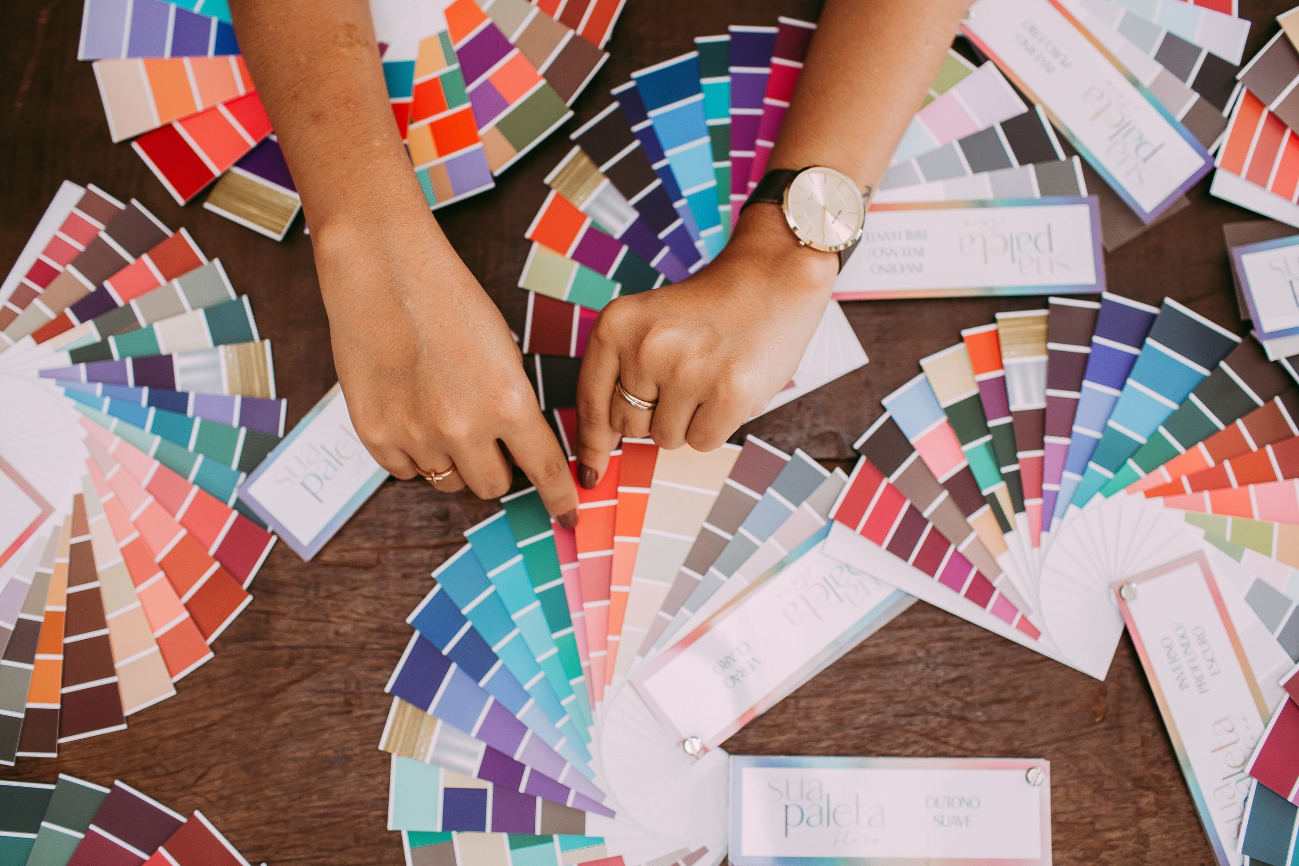 Two hands selecting colors from multiple fan decks of paint swatches spread on a wooden table.
