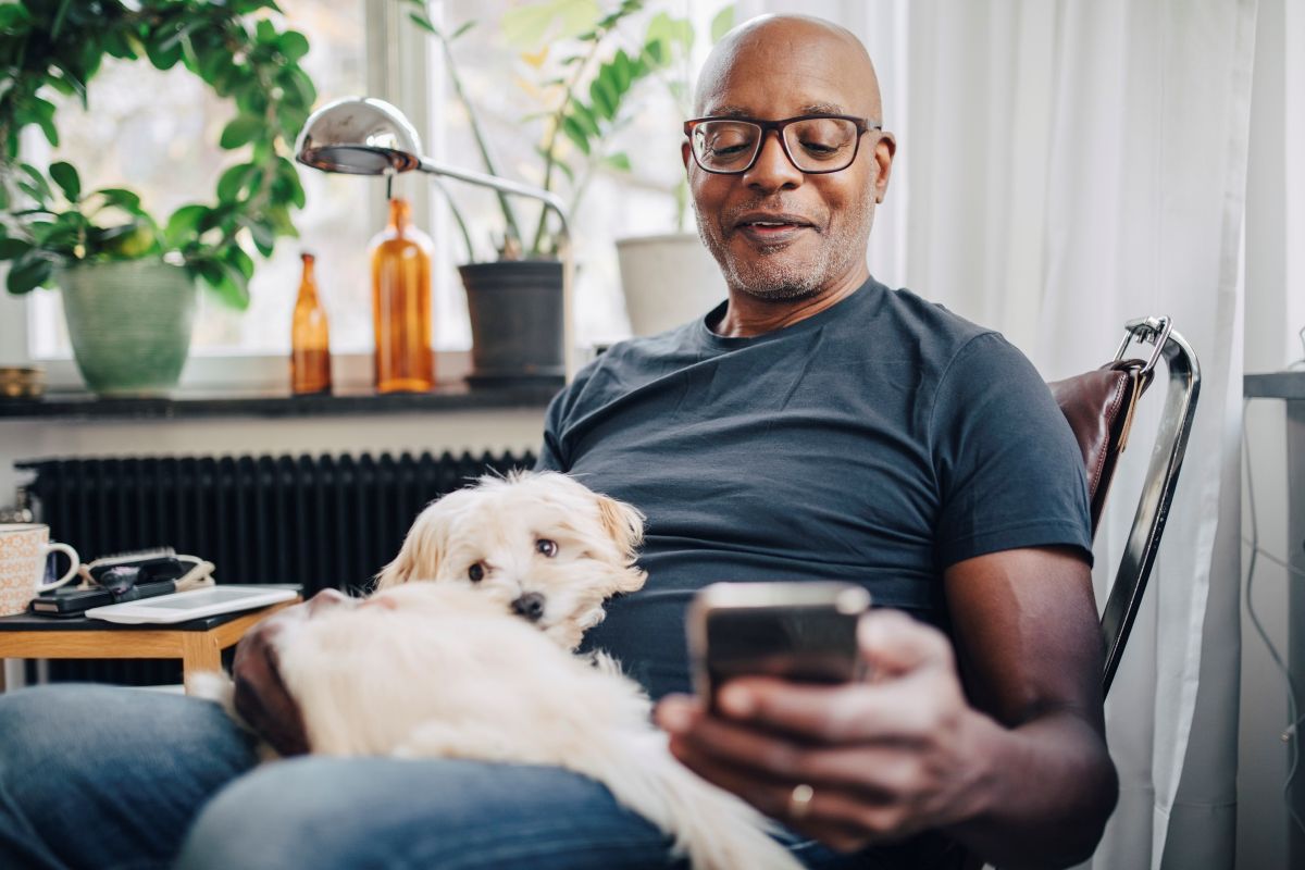 Man wearing glasses holding a smartphone with a small white dog on his lap indoors near plants.