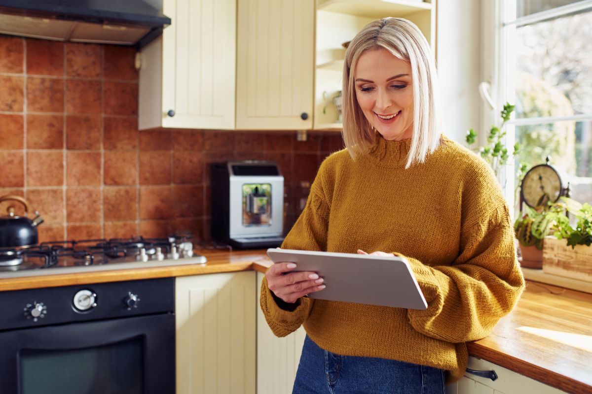 Smiling woman in mustard sweater using tablet in sunny kitchen with plants and stove behind her.