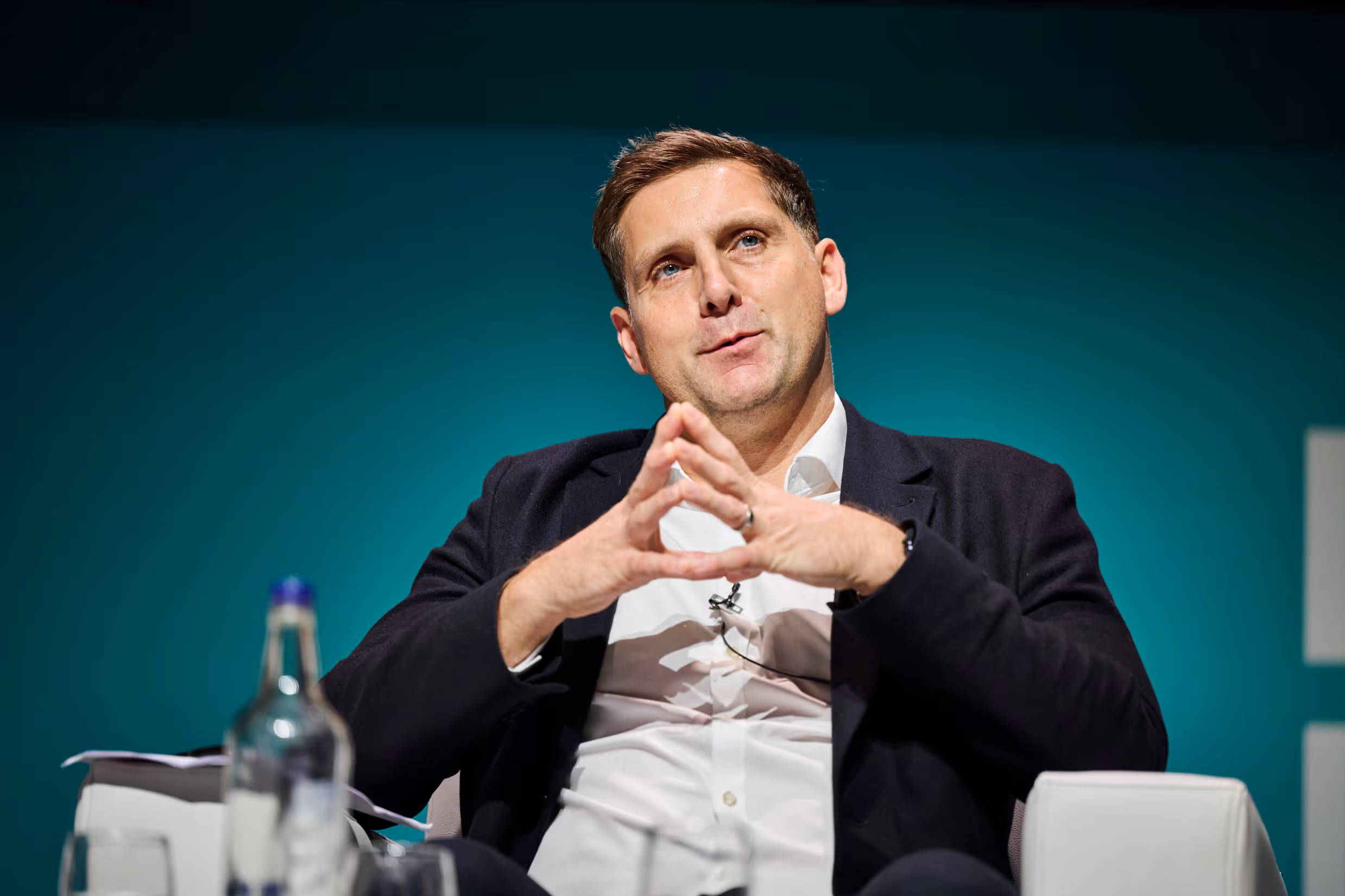 Man in white shirt and black jacket sitting with fingers steepled, speaking at event.