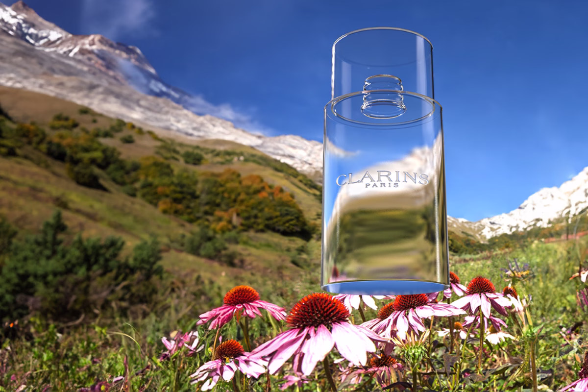 Transparent Clarins Paris bottle over pink flowers with green hills and snow-capped mountain background.