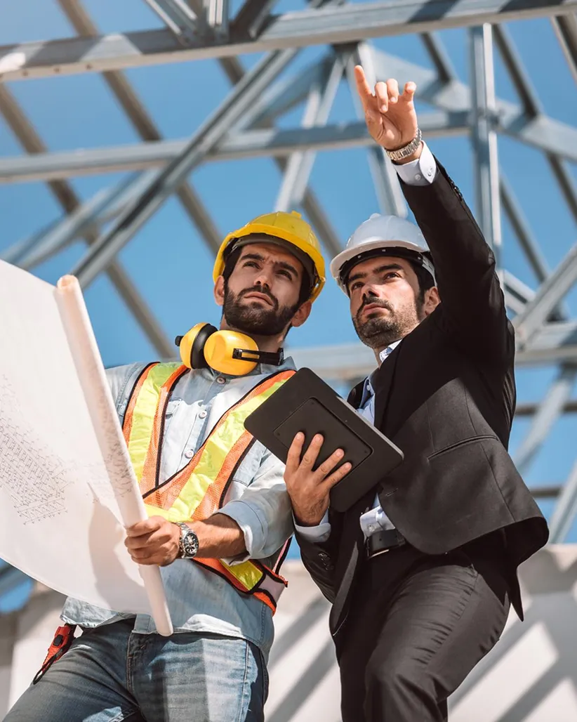 Two construction professionals, one in a yellow hard hat and safety vest holding blueprints and the other in a suit with a white hard hat holding a tablet, discussing plans at a construction site.