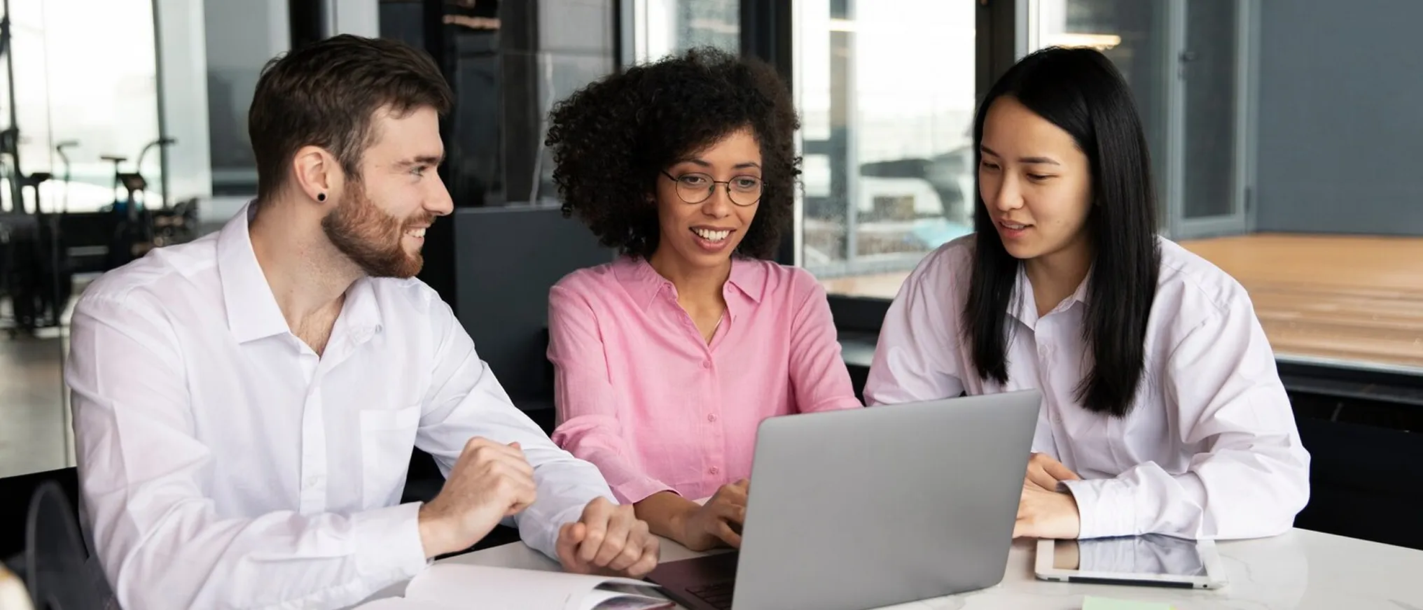 Three diverse young professionals sitting at a table, smiling and collaborating over a laptop in a modern office.