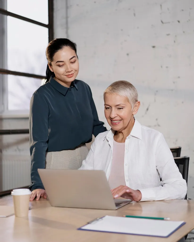 Two women working together with a laptop in an office, one sitting and the other standing beside her.