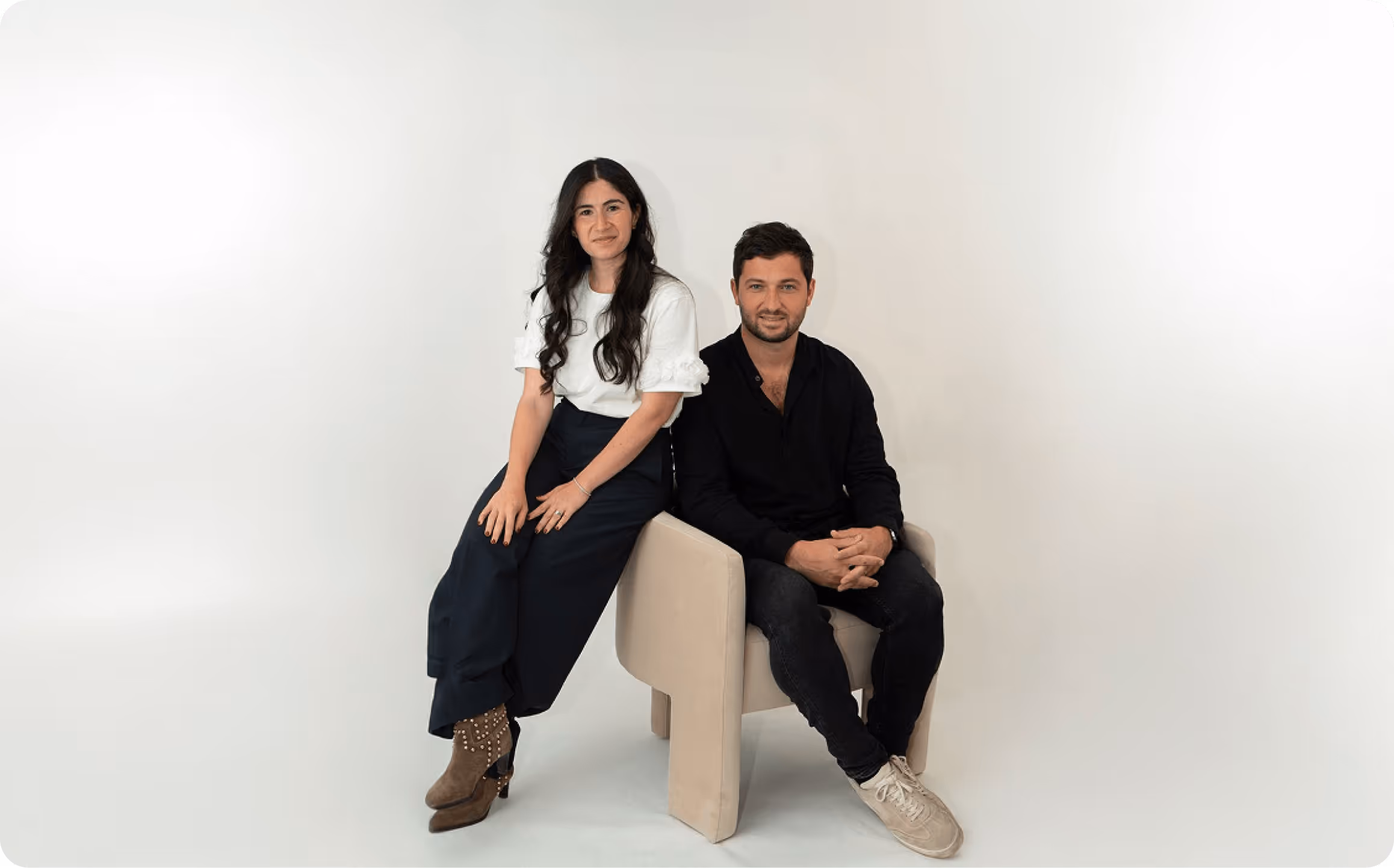 Un homme assis sur une chaise beige et une femme debout à côté de lui dans un studio avec un fond blanc.