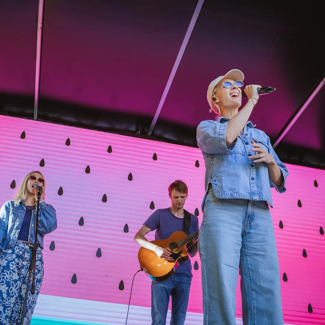 Tři hudebníci vystupující na pódiu, jedna žena zpívající do mikrofonu, oblečená v džínách a slunečních brýlích, muž hrající na akustickou kytaru a další žena zpívající v modrých květinových kalhotách před růžovo-černým pozadím.
