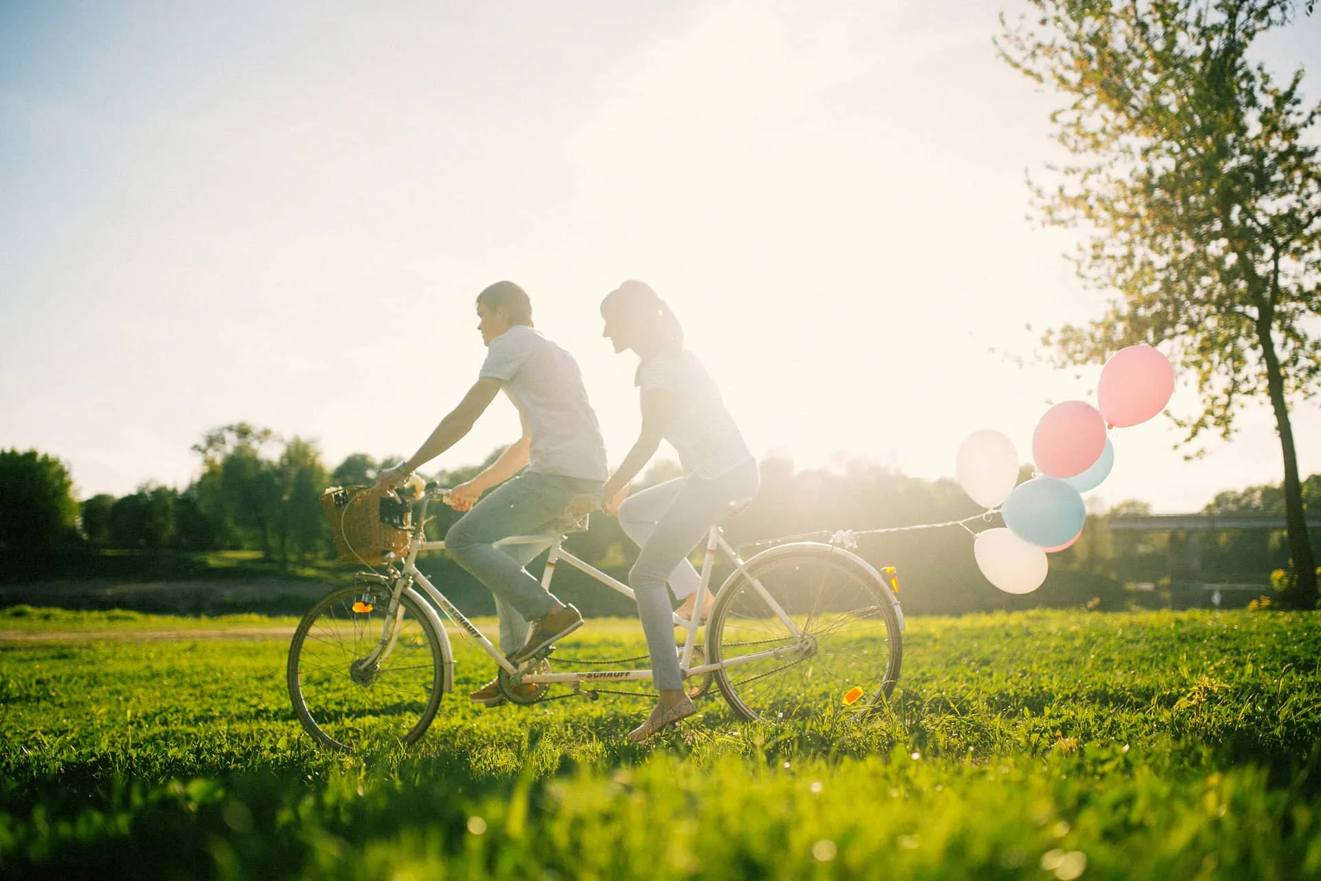 Pár jedoucí na tandemovém kole ve slunném parku s barevnými balónky přivázanými k zadní části.