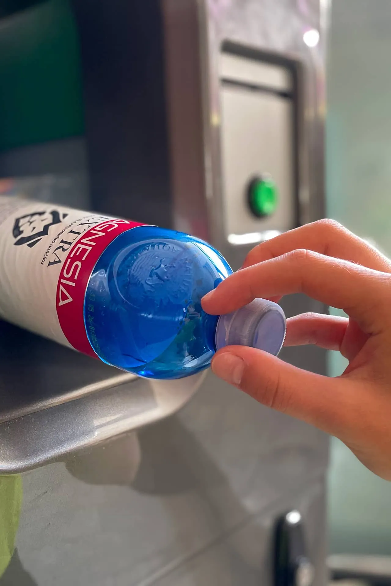 Ruka držící víčko modré plastové láhve s vodou s nápisem Magnesia položenou na kovovém povrchu kontejneru pro recyklaci.