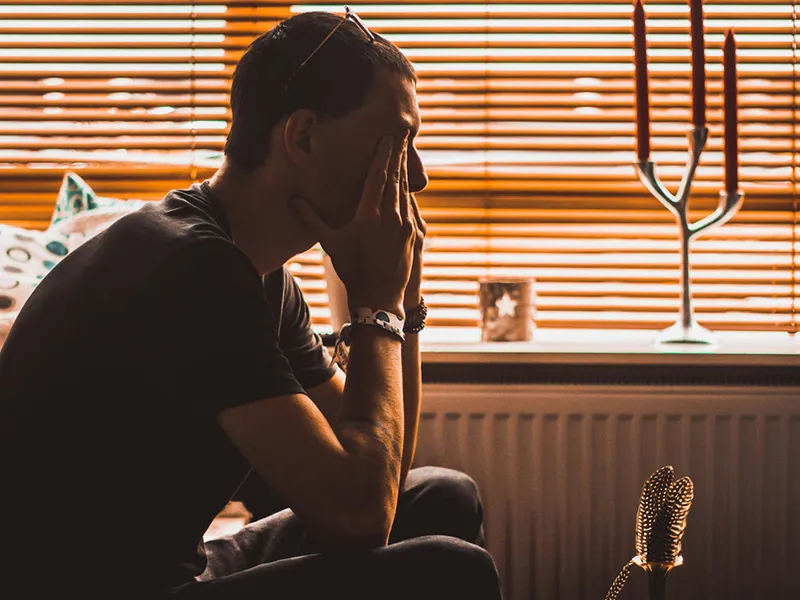 Muž sedící s hlavou v dlaních v tlumeně osvětlené místnosti s roletami a svíčkami v pozadí.