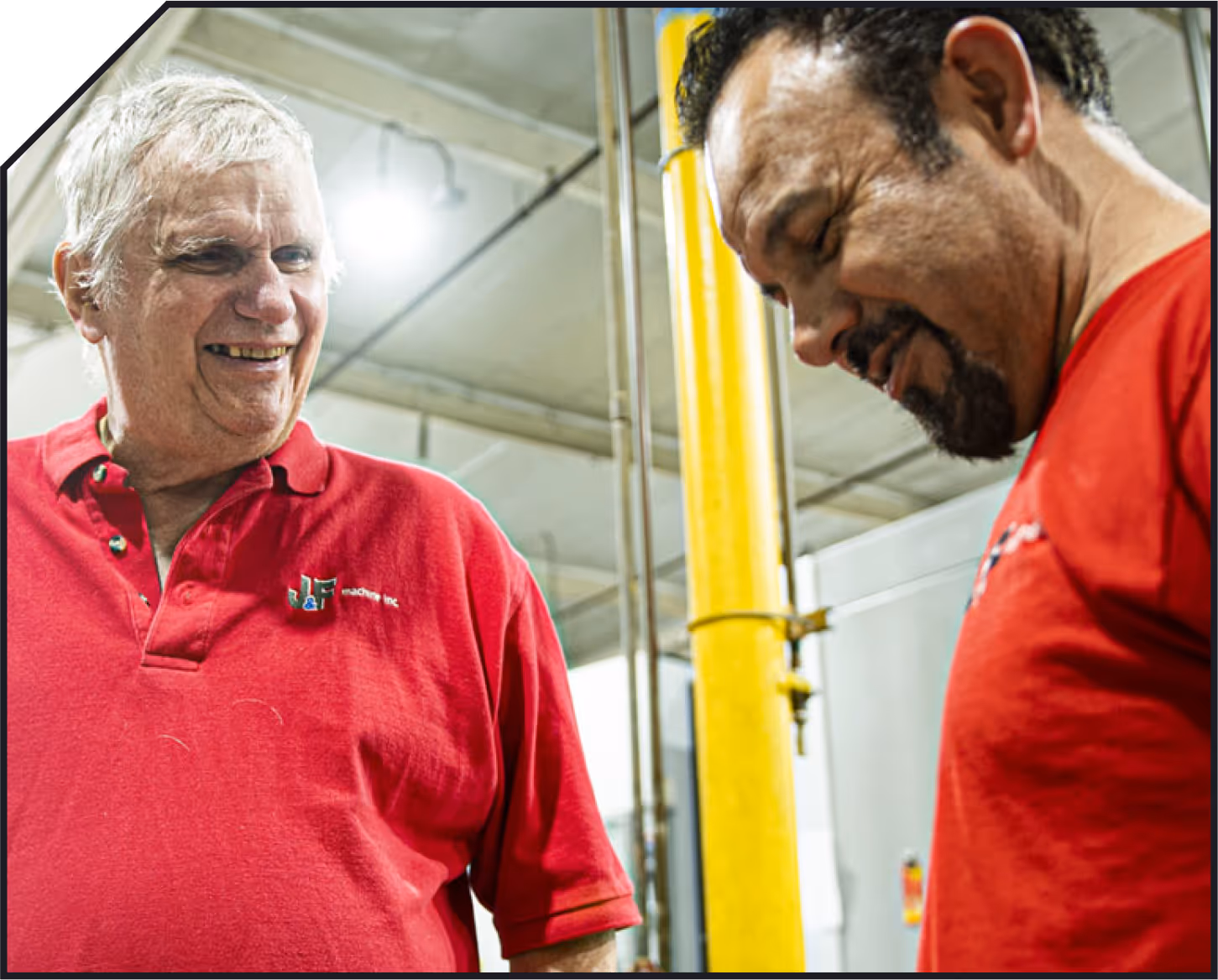 Two men in red shirts smiling and interacting in an industrial setting with a yellow pillar in the background.
