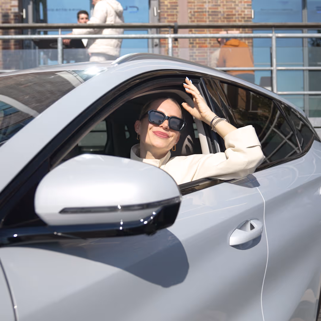 Frau in Sonnenbrille im weißen Auto lächelt und hebt die Hand zum Haarekämmen.