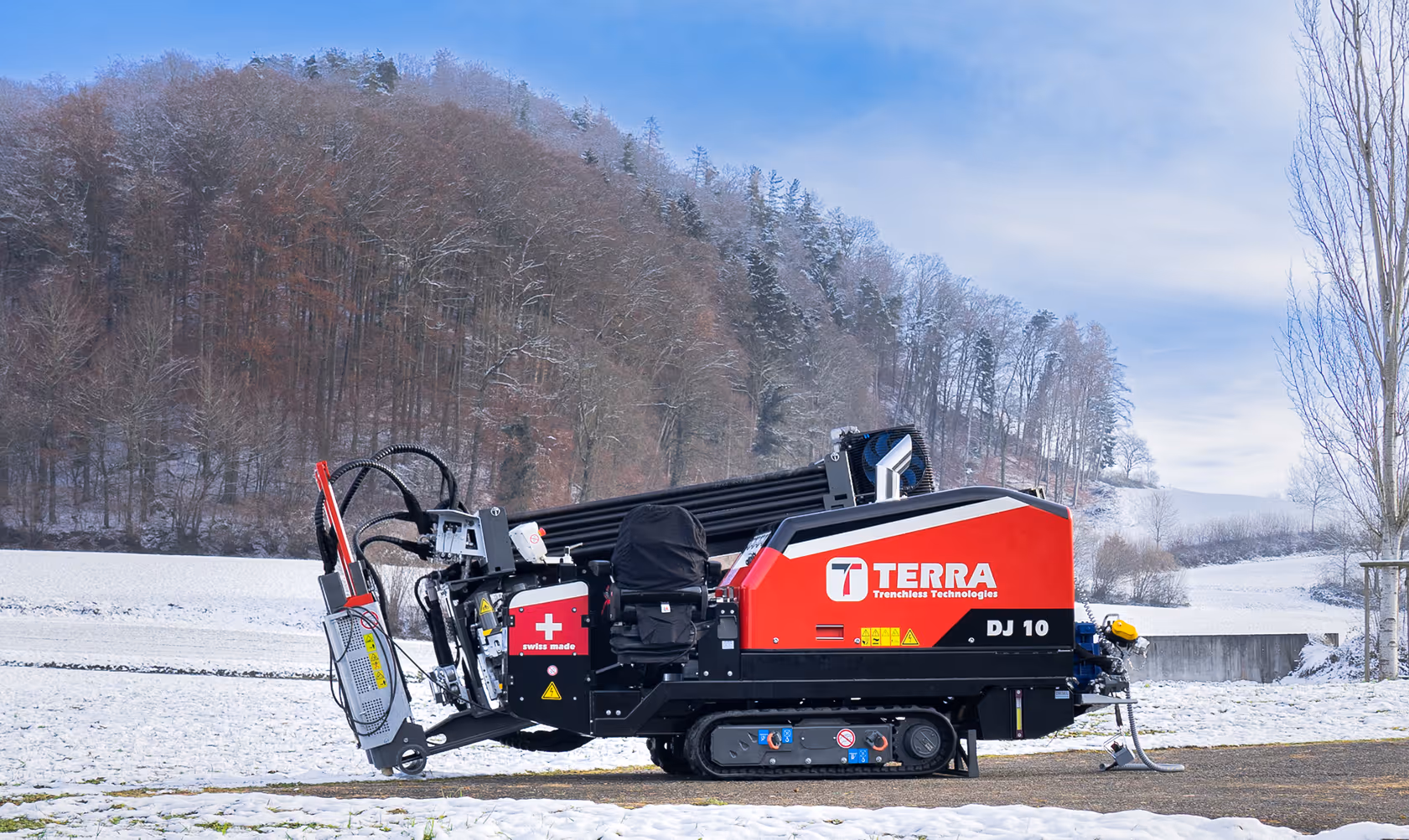 Rotes und schwarzes Terra DJ 10 Grabenlos-Bohrgerät auf Schnee vor bewaldetem Hügel bei klarem Himmel.