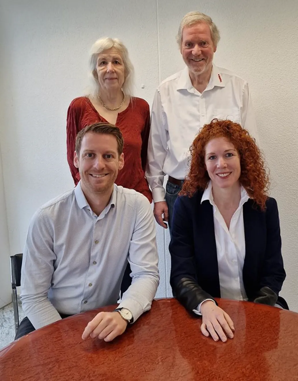 Quatre personnes, deux femmes et deux hommes, sourient dans un bureau et s'assoient ou se tiennent debout autour d'une table ronde en bois.