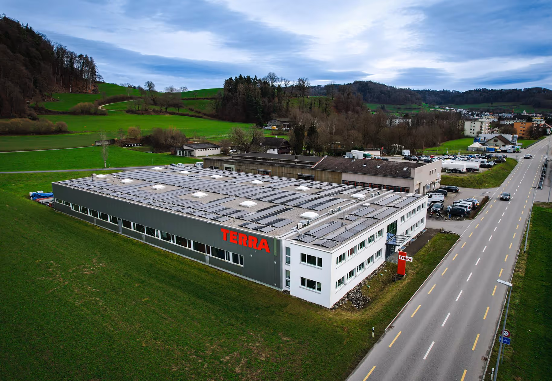 Vue aérienne d'un grand bâtiment de bureaux et de production avec des panneaux solaires sur le toit dans une zone rurale verdoyante à côté d'une route.