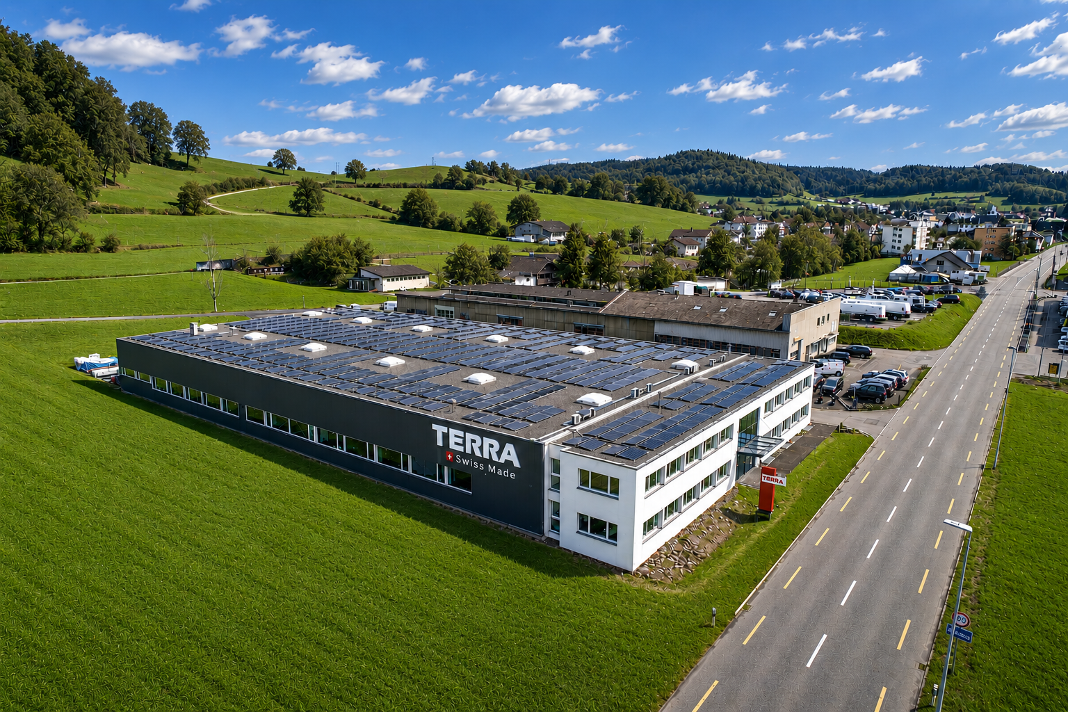 Luftaufnahme eines grossen Büro- und Produktionsgebäudes mit Solarzellen auf dem Dach in grüner ländlicher Umgebung neben einer Strasse.