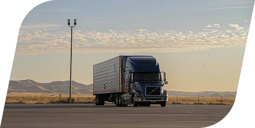 Black semi-truck parked on an empty lot with a backdrop of mountains and a cloudy sky at sunset.