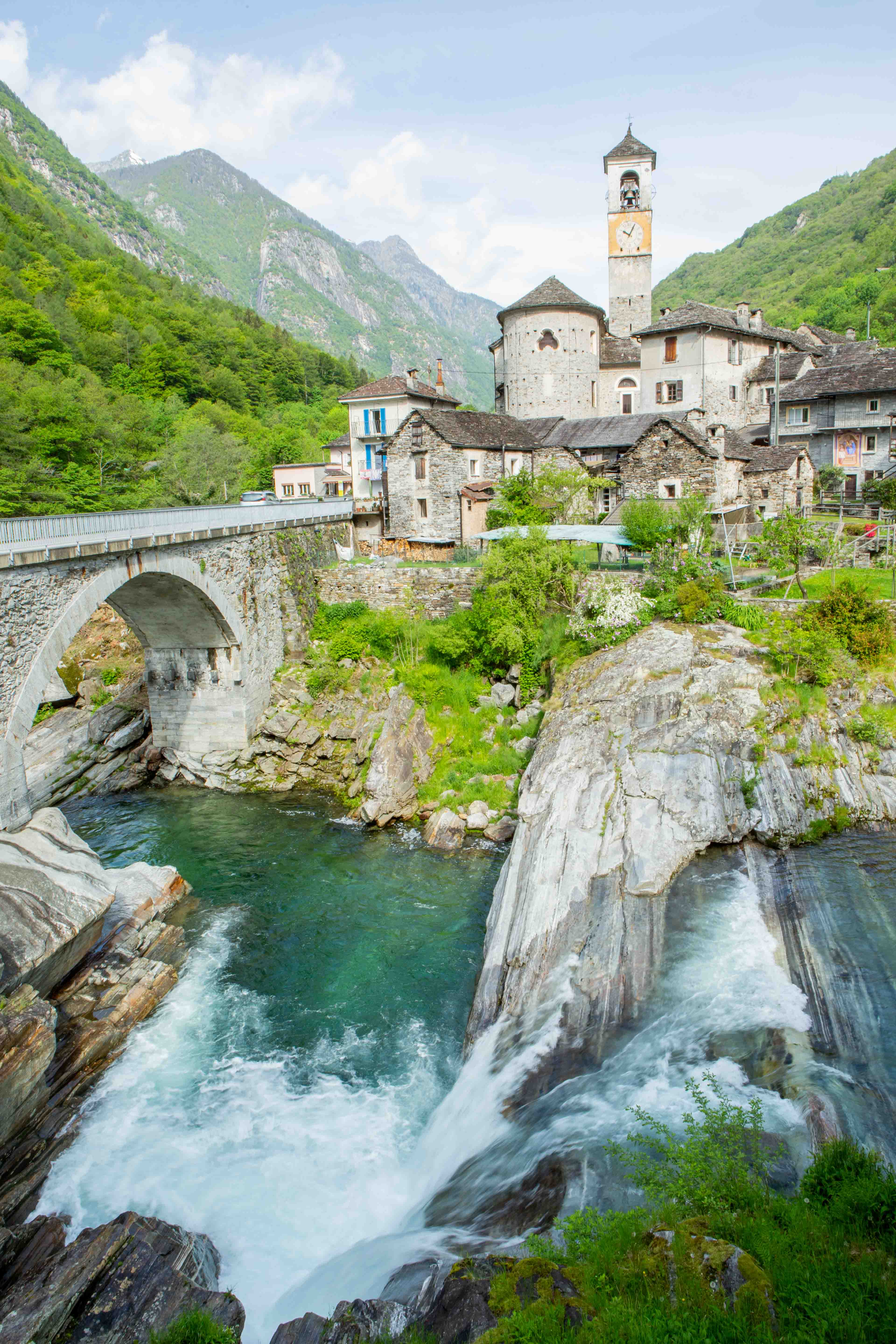 Stone bridge over rushing river beside stone buildings with a bell tower in a green mountain valley.
