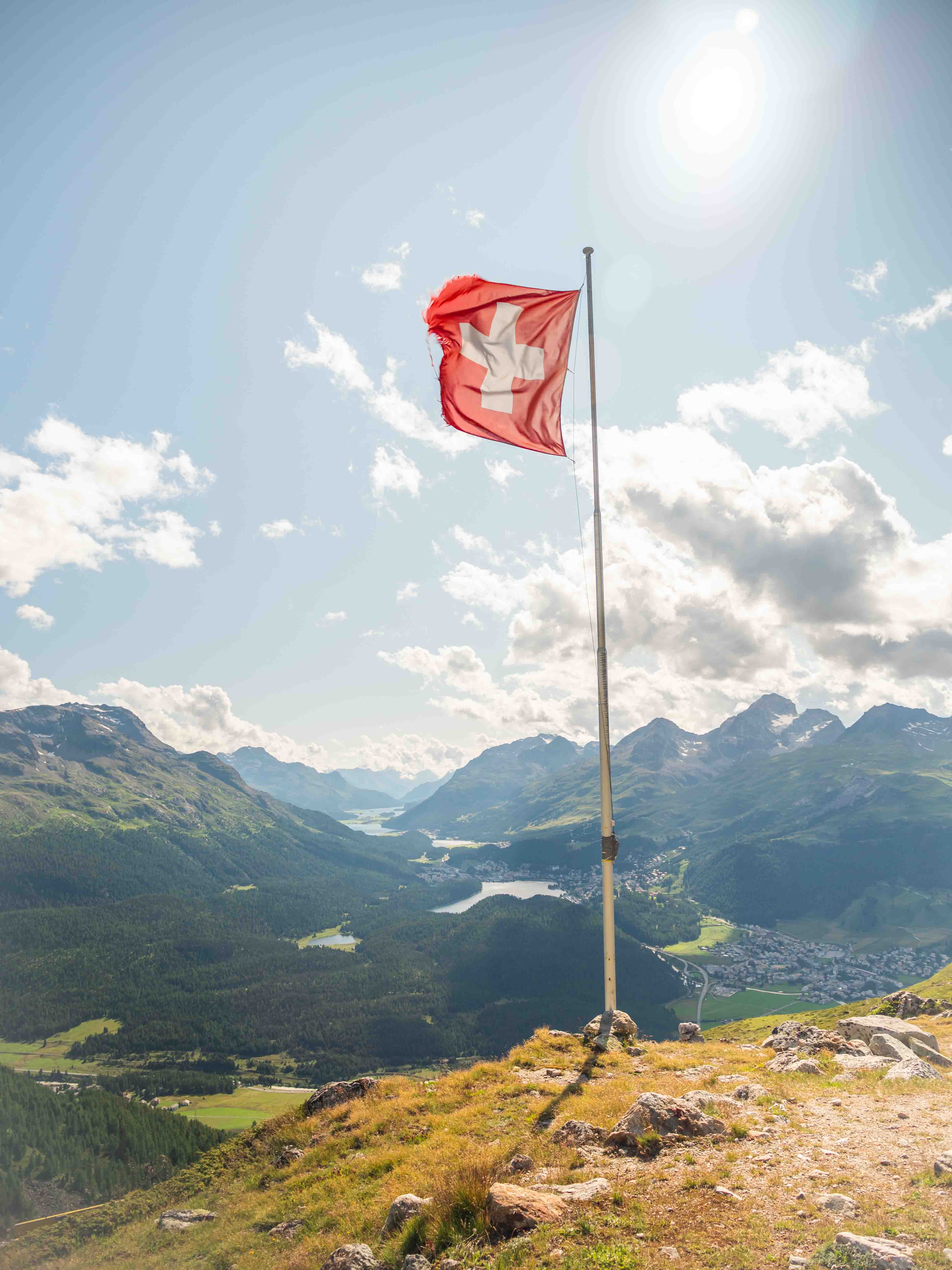 Swiss flag waving on a pole atop a grassy mountain overlooking a valley and distant peaks.