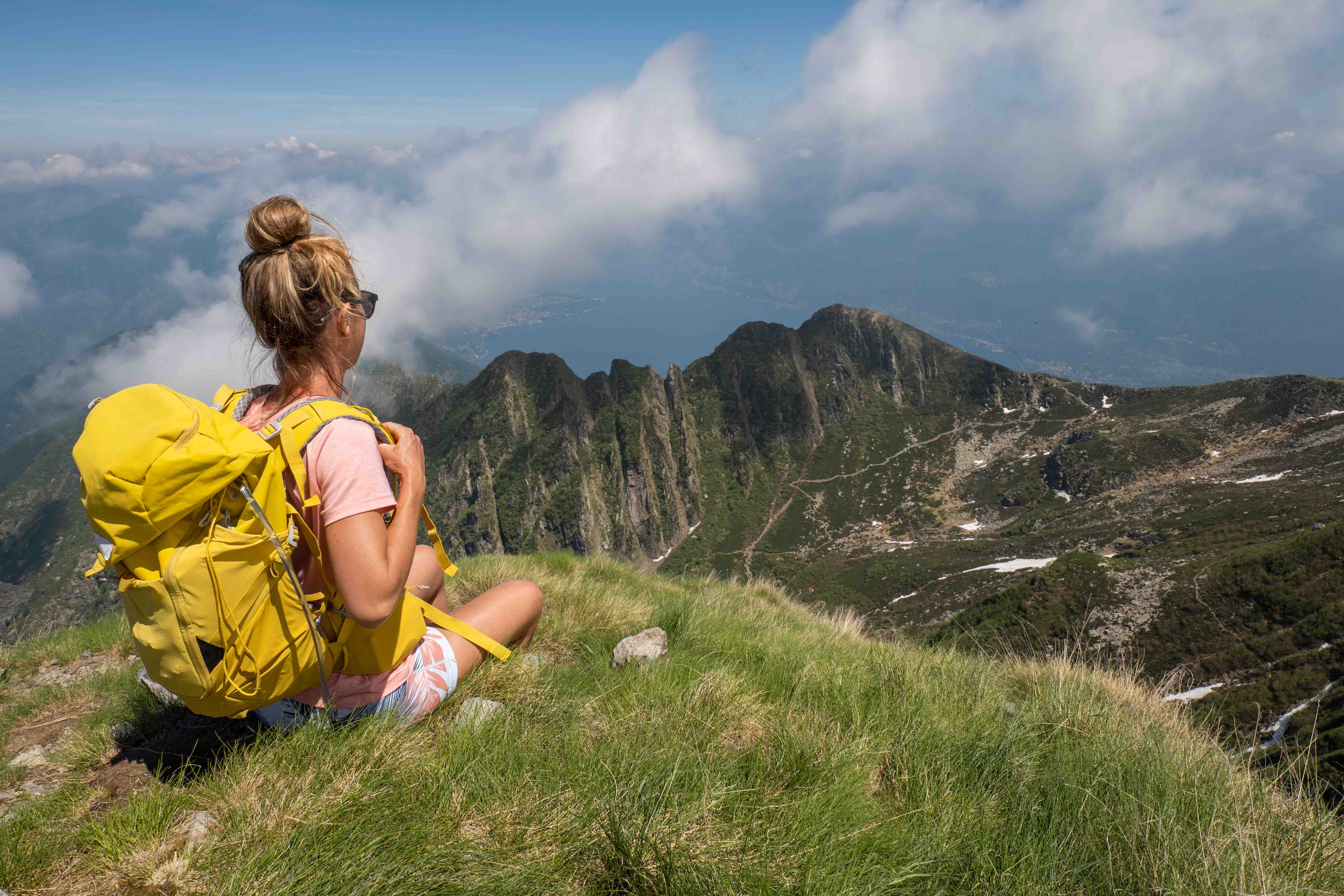 Woman with yellow backpack sitting on grassy mountain ledge overlooking peaks and cloudy sky