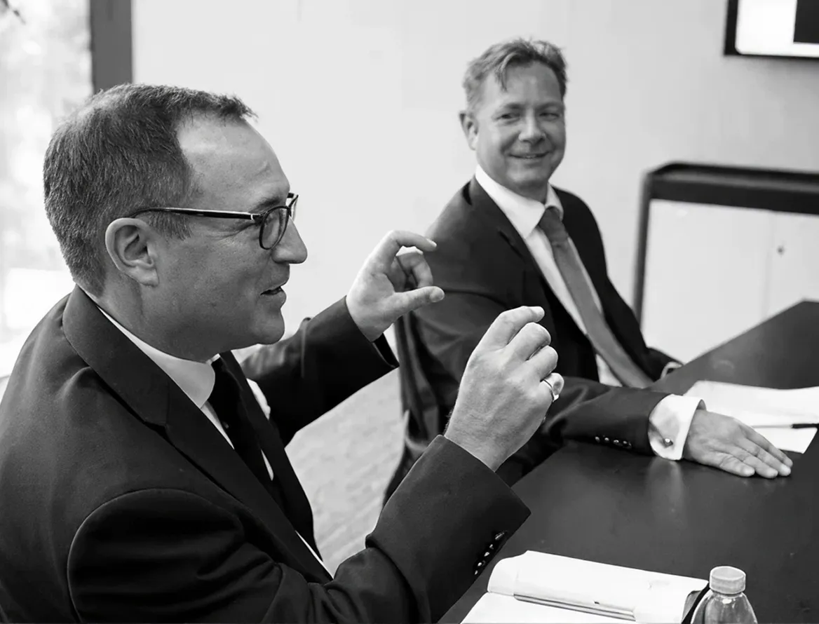 Two men in suits having a discussion at a conference table, one gesturing with his hands while the other listens with a smile.