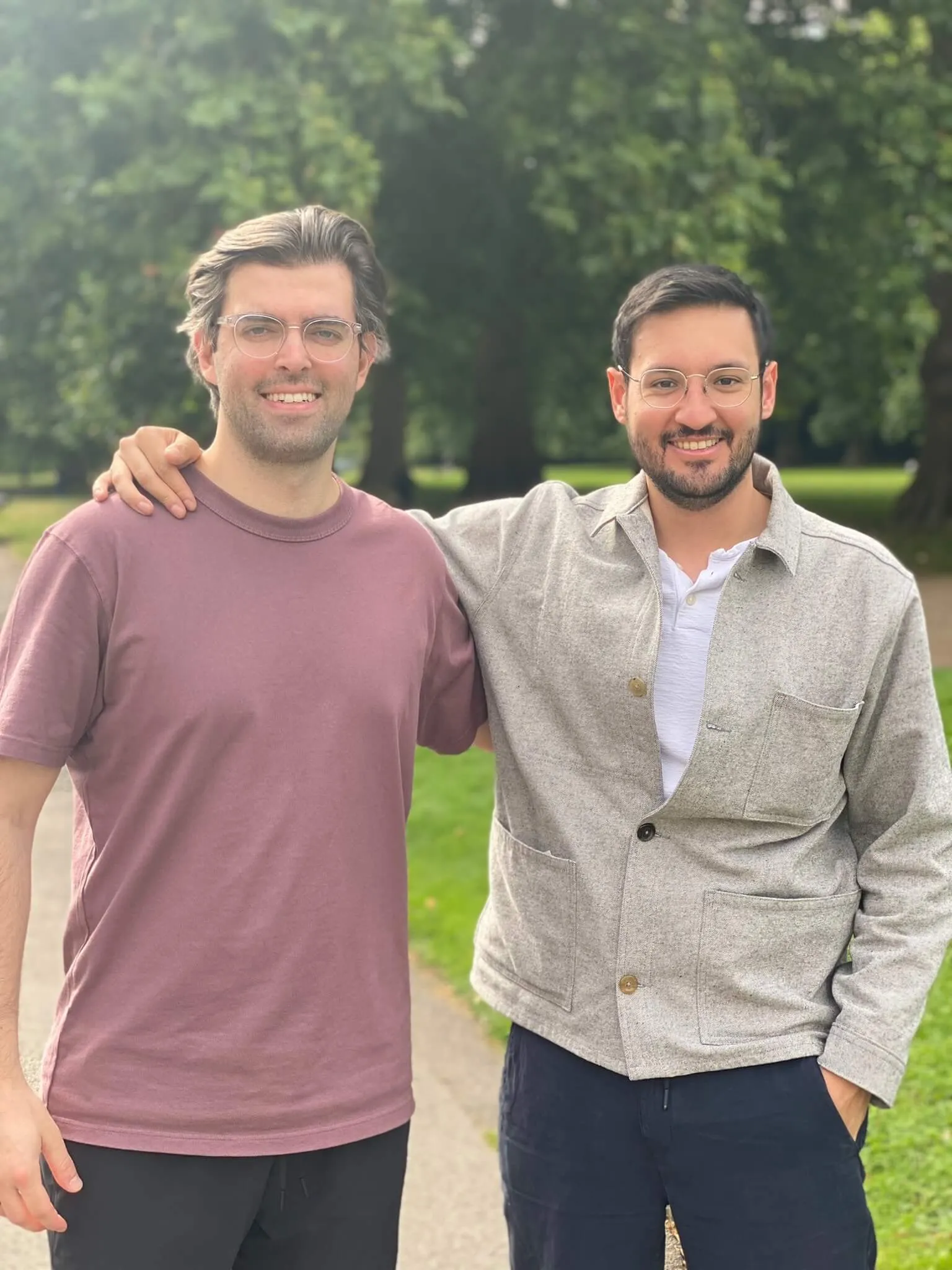 Two men wearing glasses stand side by side outdoors with arms around each other, smiling.