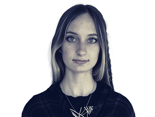 Young woman with light hair and blue eyes staring straight, wearing a dark shirt and necklace.