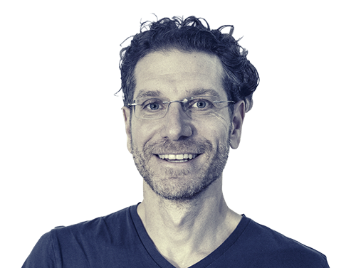 Smiling man with curly dark hair, glasses, and facial stubble wearing a black V-neck shirt.