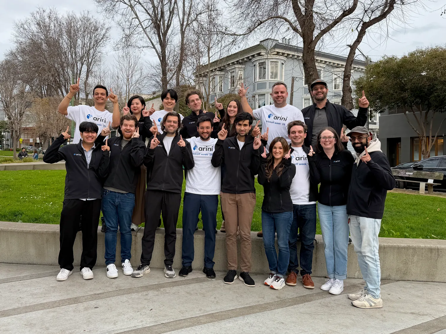 Group of 16 smiling people outdoors, some in white and black arini branded shirts, all raising one finger.