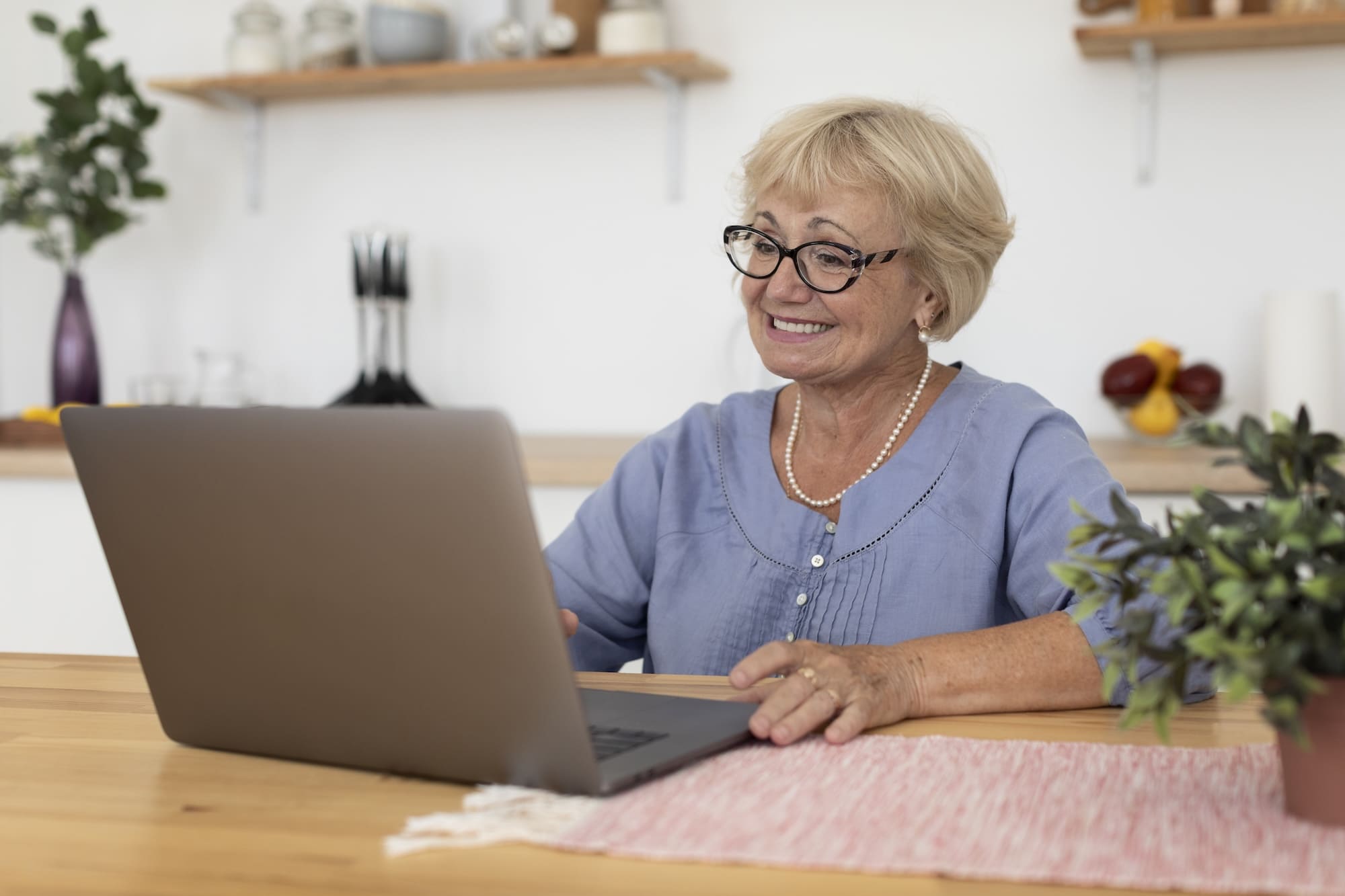 Smiling elderly woman wearing glasses and a blue blouse using a laptop at a wooden table in a kitchen.