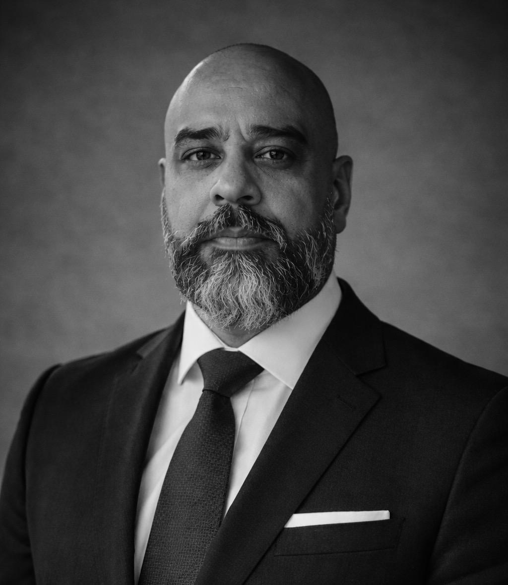 Serious bald man with a grey beard wearing a dark suit, white shirt, and tie against a plain background.