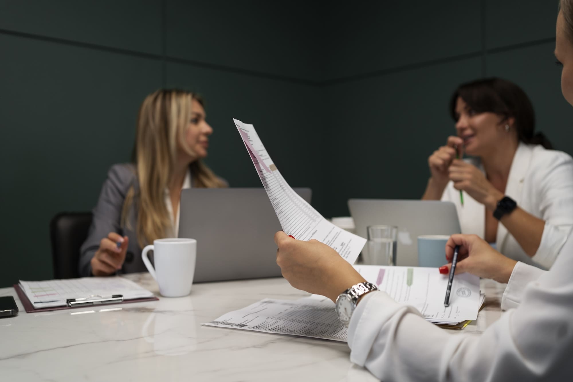Three women in a meeting room reviewing documents and working on laptops at a marble table.