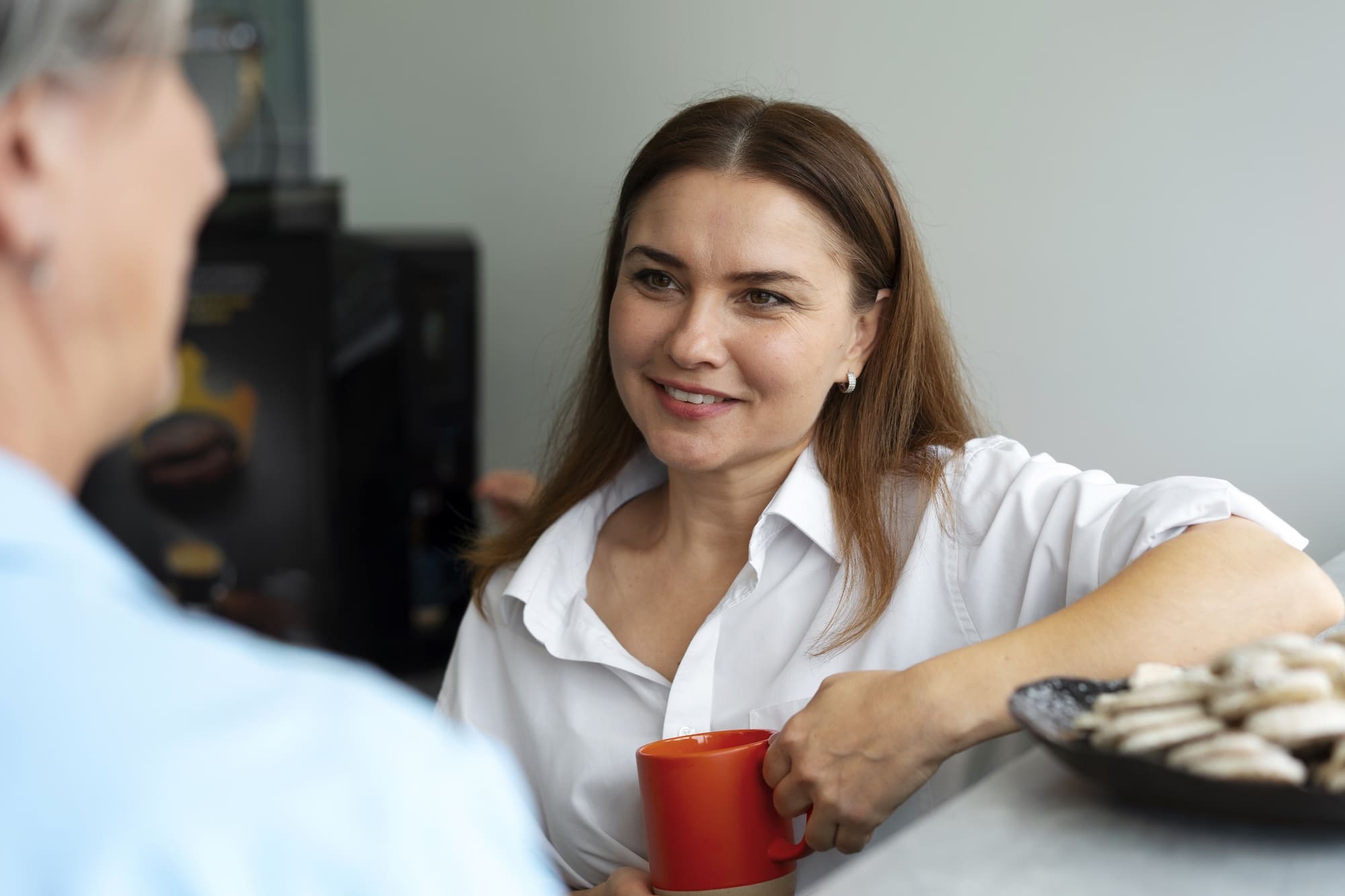 Smiling woman in white shirt holding a red mug, engaging in conversation with another person.