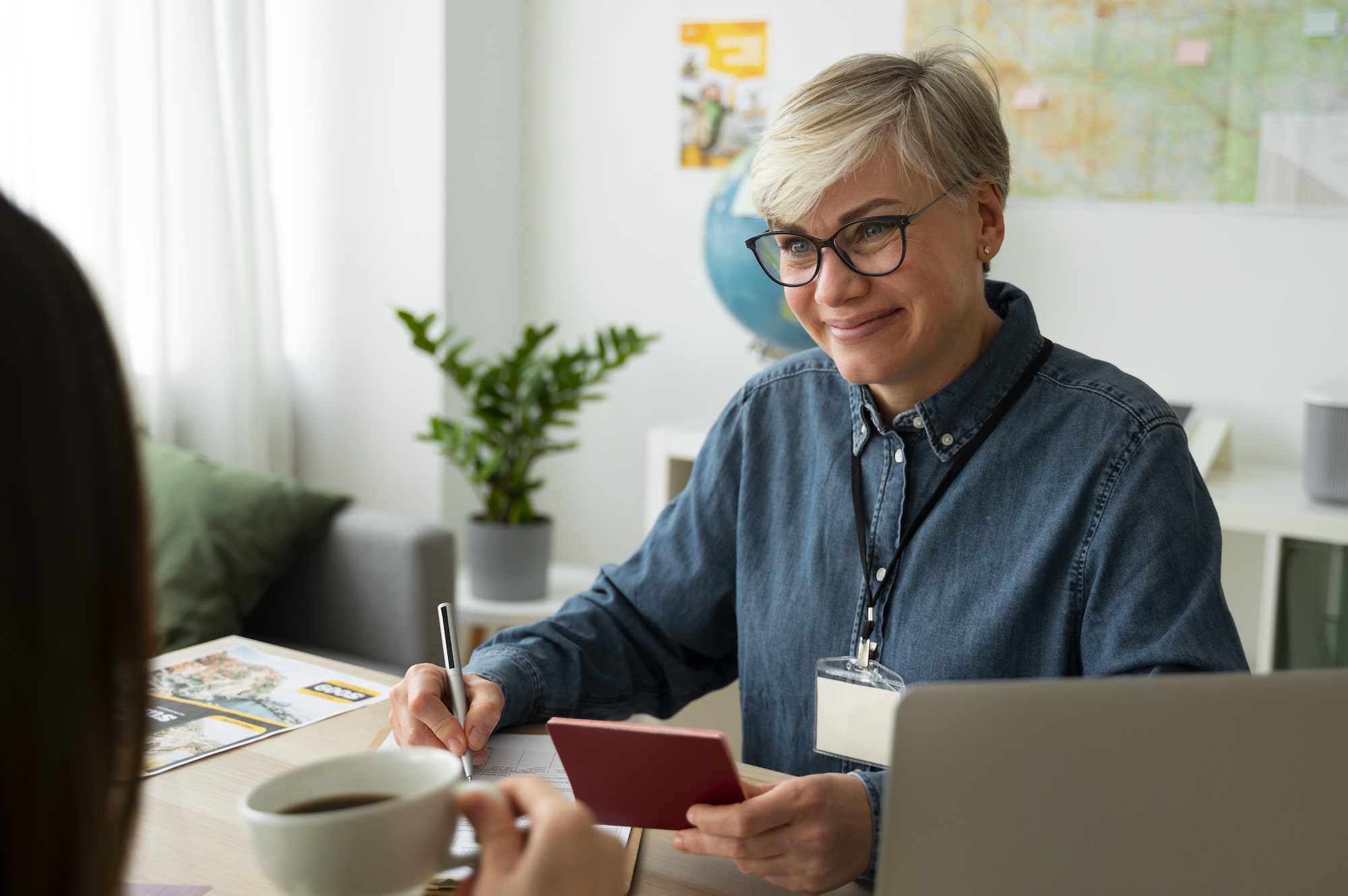 A smiling woman with glasses and short blonde hair holding a passport and pen, helping another person across a table in a bright office.
