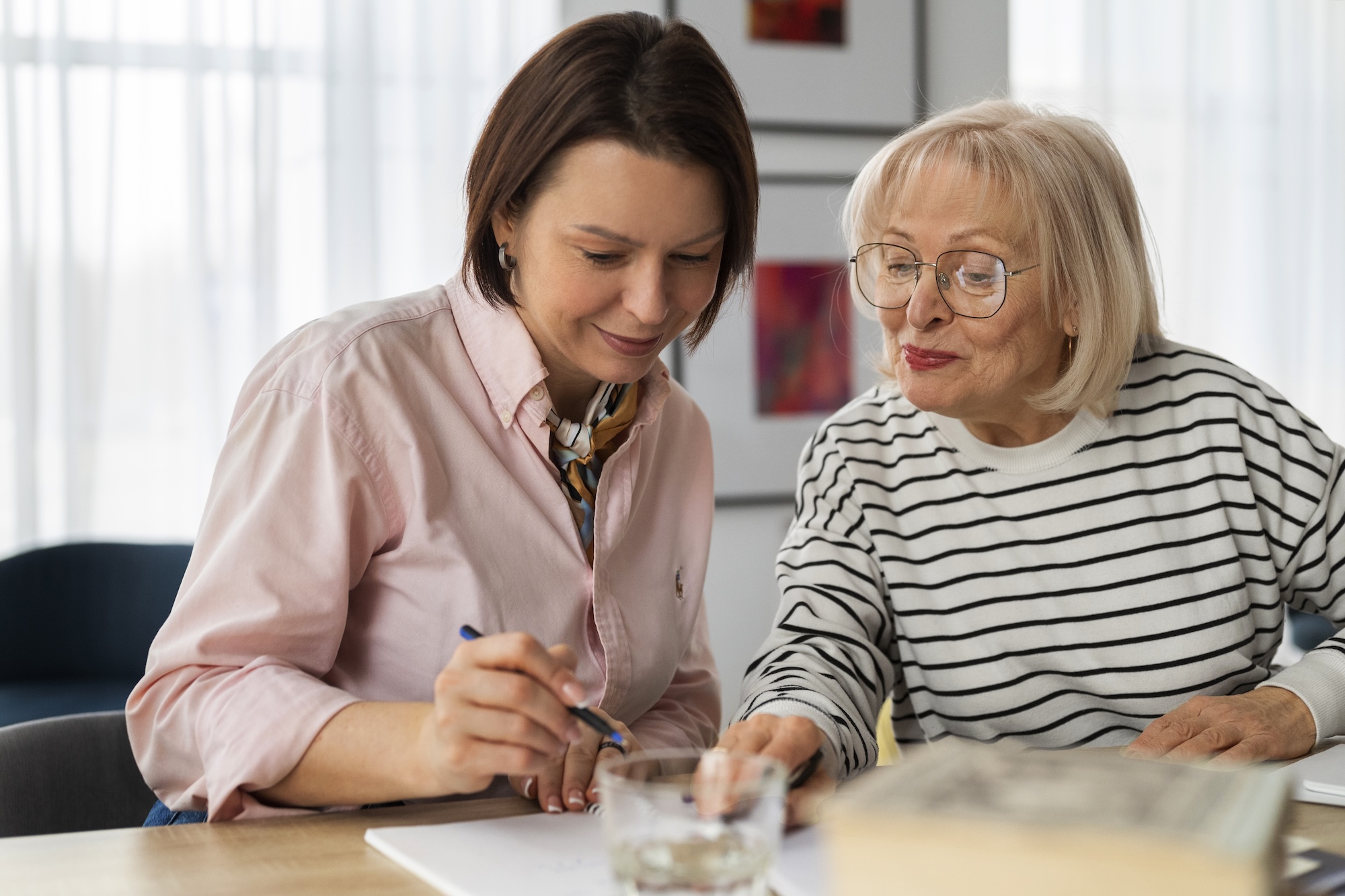 A younger woman in a pink shirt helps an older woman in a striped sweater with writing on paper at a table.