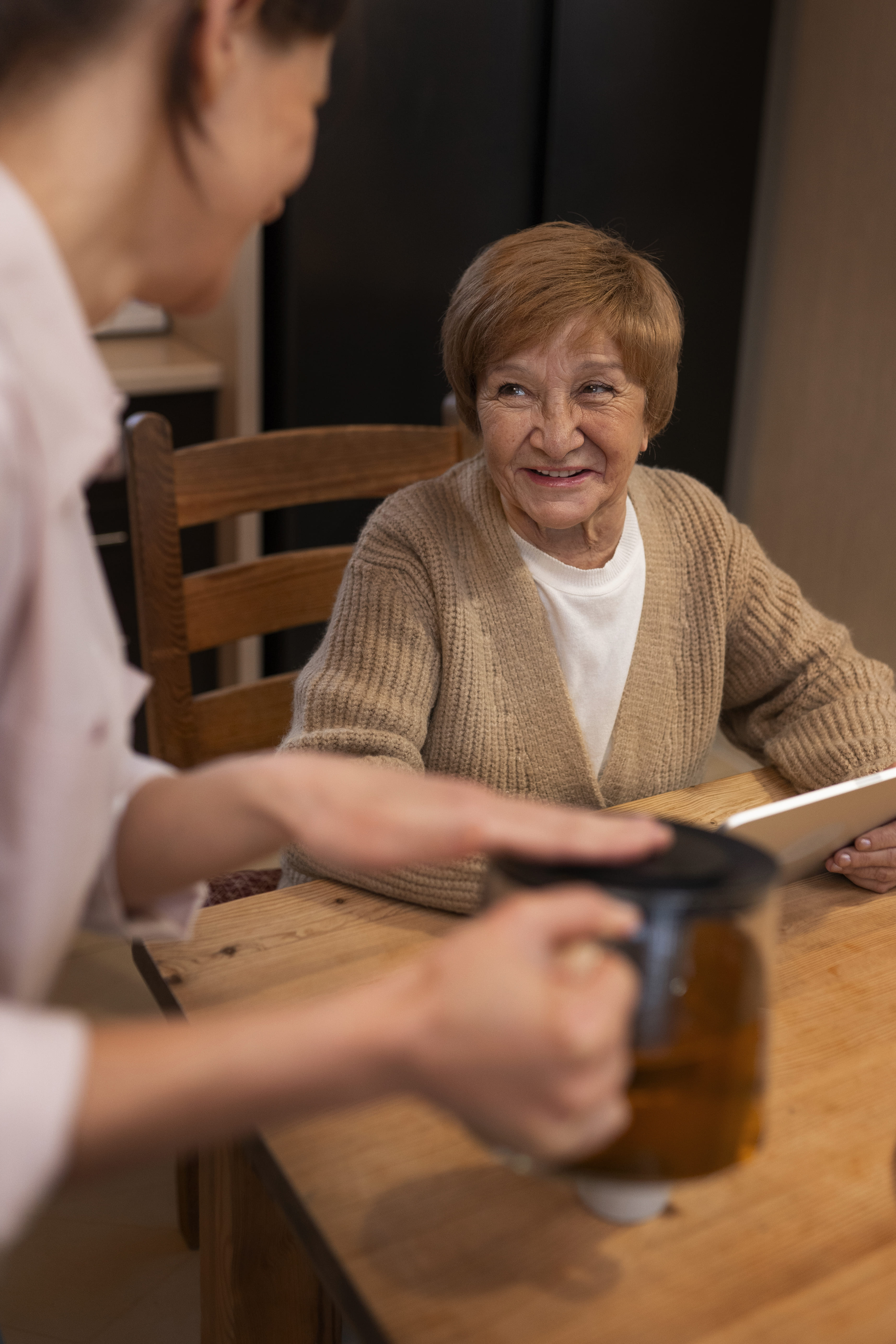 Smiling elderly woman in a beige sweater sitting at a wooden table holding a tablet, with a person in the foreground pouring tea from a teapot.