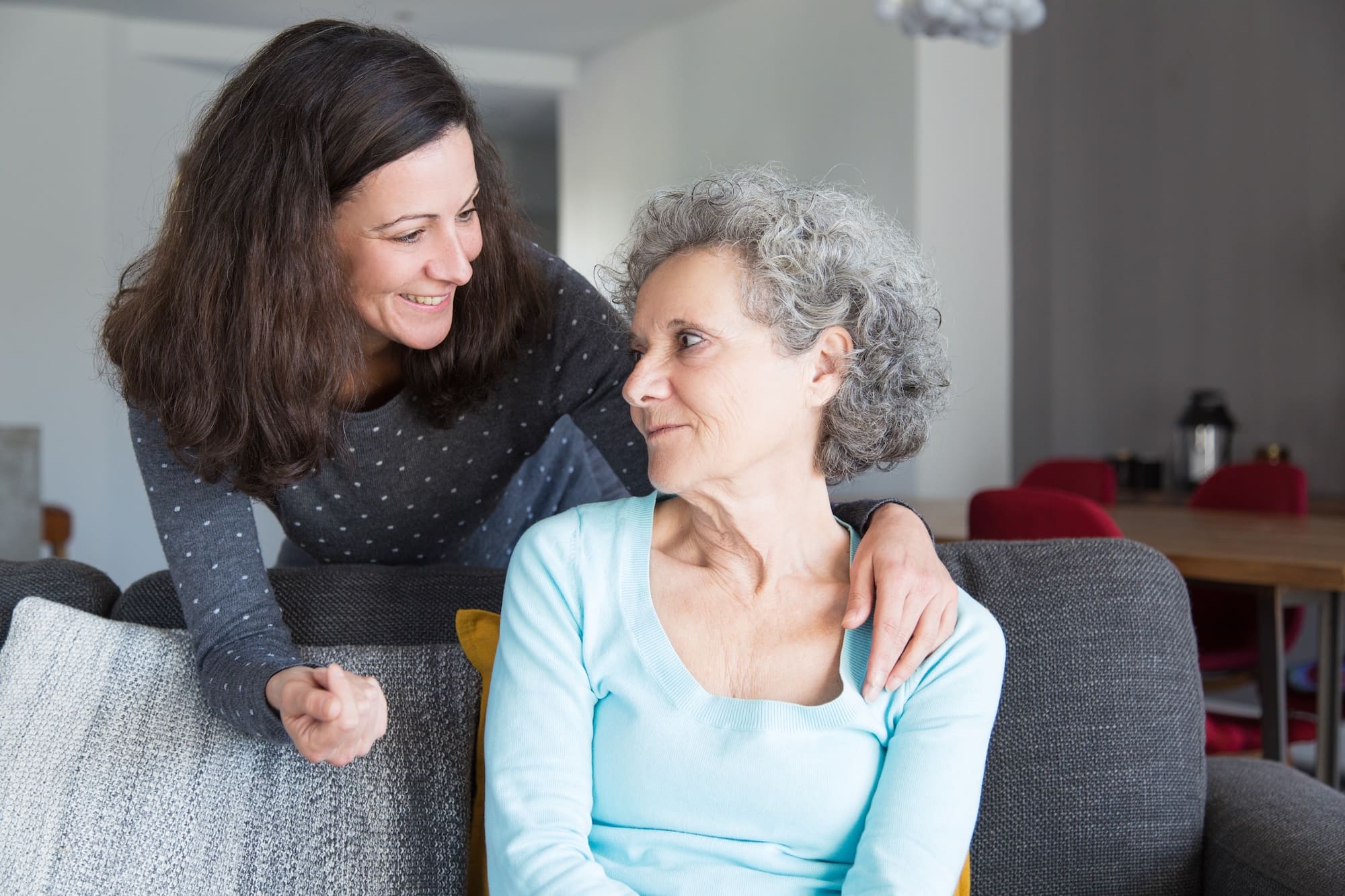 Smiling young woman with brown hair comforting an older woman with curly gray hair sitting on a couch.