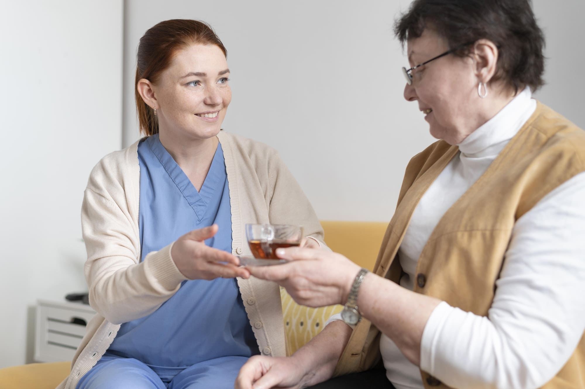 A female caregiver in blue scrubs and a beige cardigan hands a glass cup of tea to a smiling elderly woman wearing glasses and a brown vest.