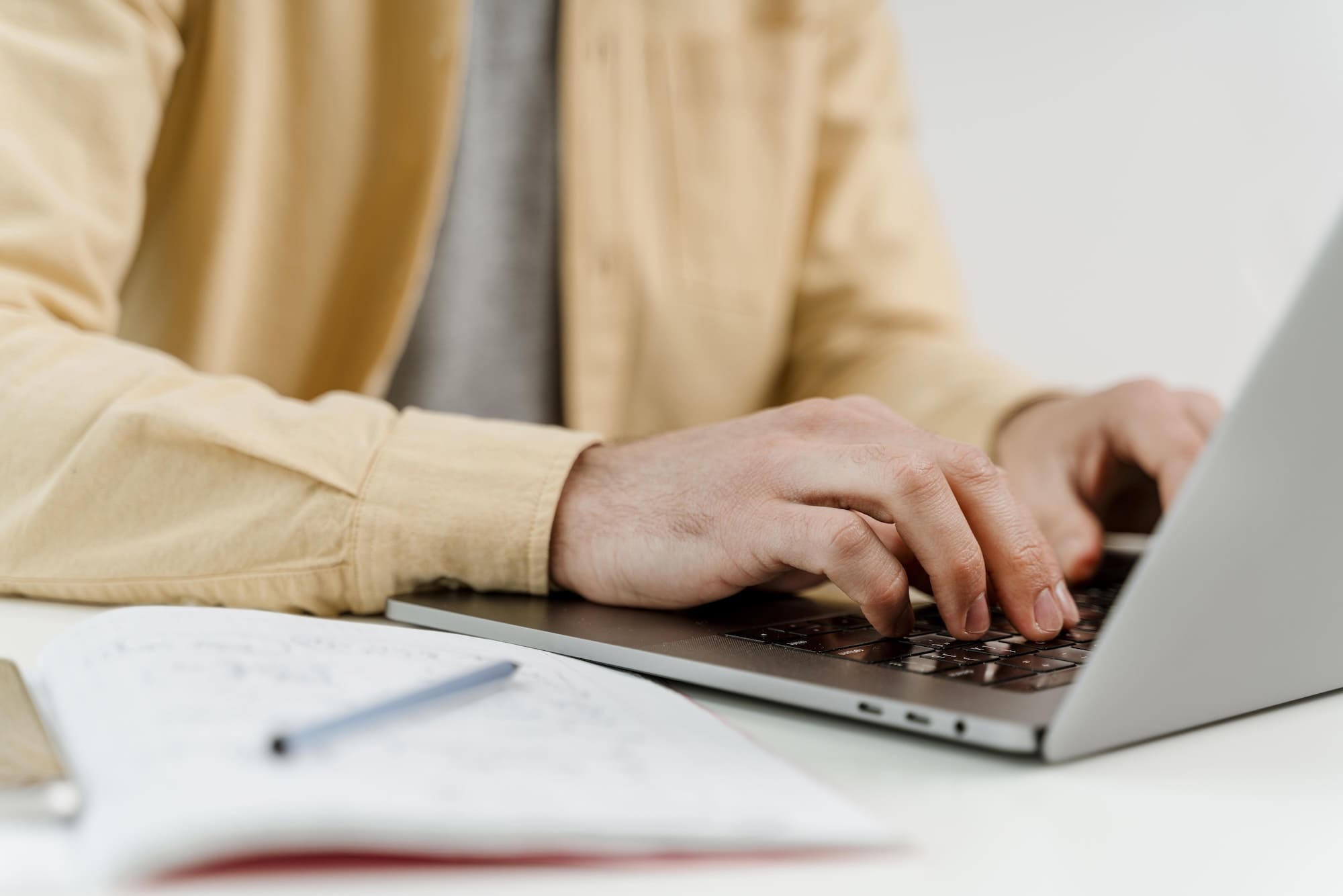 Person wearing a beige shirt typing on a laptop with an open notebook and pen on a white desk.