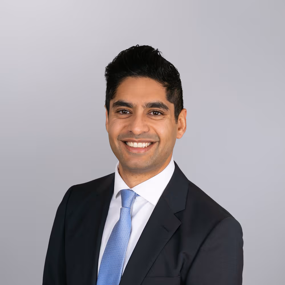 Smiling man in black suit, white shirt, and blue tie against a gray background.