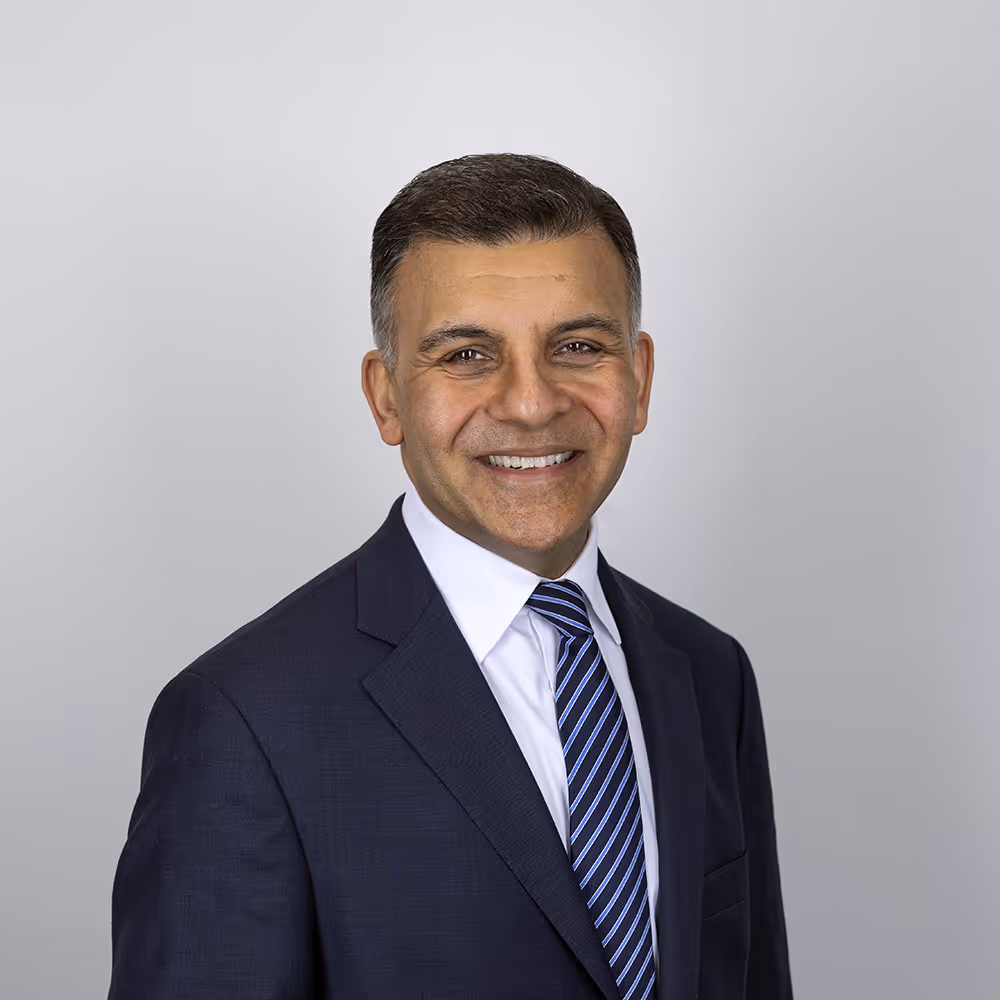 Smiling man in a dark suit, white shirt, and blue striped tie against a light gray background.