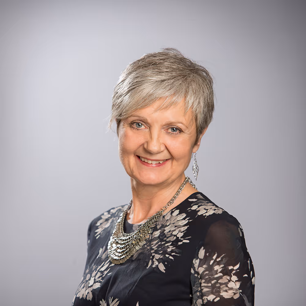 Smiling woman with short gray hair wearing a dark floral top and silver necklace and earrings against gray background.